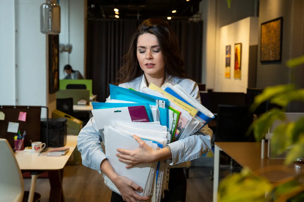 Femme en train de porter des piles de chemises dossier et de classeur encourant une chute faisant partie des risques du métier archiviste